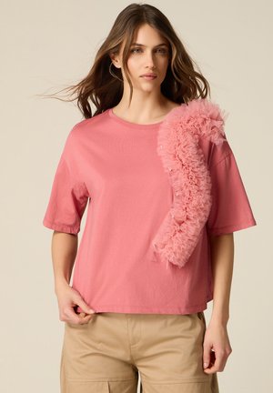 Donna che indossa un top rosa a maniche corte con un grande dettaglio in tulle arricciato su una spalla e pantaloni beige, in piedi davanti a uno sfondo semplice.