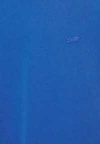 Tissu bleu avec un petit logo cousu en fil bleu assorti près du centre à droite.