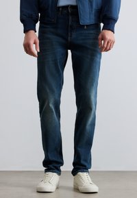 Σκούρο μπλε τζιν παντελόνι με στενή γραμμή, με ξεθωριασμένο πλύσιμο και διακριτικές γραμμές φθοράς, συνδυασμένο με λευκά χαμηλά αθλητικά παπούτσια.