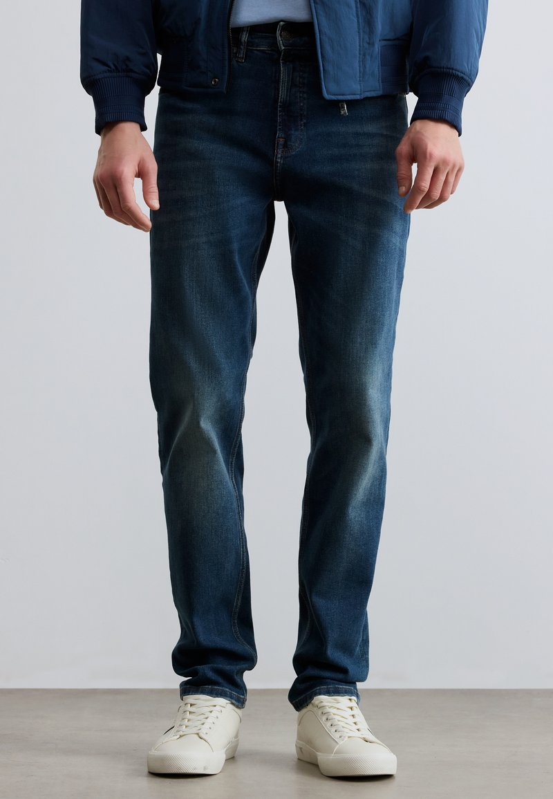 Σκούρο μπλε τζιν παντελόνι με στενή γραμμή, με ξεθωριασμένο πλύσιμο και διακριτικές γραμμές φθοράς, συνδυασμένο με λευκά χαμηλά αθλητικά παπούτσια.