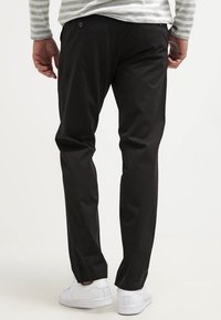 Black, slim-fit pants made from smooth fabric, featuring back pockets and a button accent, paired with white sneakers.