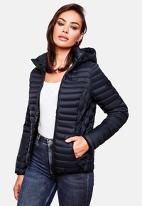 Black puffer jacket with a hood, featuring quilted texture, zippered pockets, and a slim fit. Worn over a white top and denim jeans.