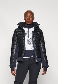 Giacca imbottita nera e blu con zip frontale, maniche spesse e cappuccio. Presenta una maglietta bianca grafica sotto e pantaloni scuri aderenti.