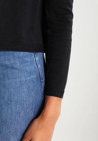 Top negro de manga larga con dobladillo redondeado, combinado con unos jeans de mezclilla azules. La tela parece suave con una ligera textura.
