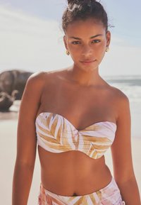 Bikini-top heeft een strapless ontwerp met een witte achtergrond en gouden palmblads patronen. Glad stof met subtiele textuur en een geplooid middenstuk.