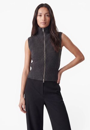 Femme aux longs cheveux foncés portant un gilet en maille gris foncé sans manches avec fermeture éclair et un pantalon noir taille haute, debout avec une main sur la hanche.