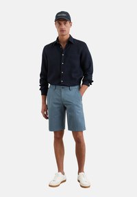 Donkerblauw overhemd met lange mouwen en knoopsluiting, lichtblauwe shorts en witte sneakers met tan zolen. Pet met logo-tekst.