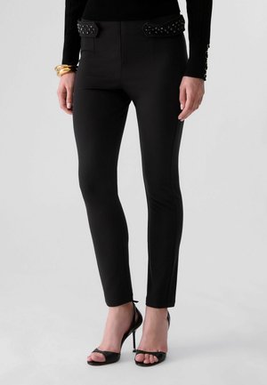 Femme portant un pantalon noir ajusté avec des accents décoratifs cloutés et des talons hauts noirs ouverts à l’avant, debout avec les mains détendues le long du corps.