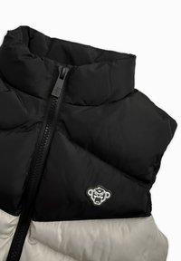 Schwarze und cremefarbene Steppjacke mit hohem Kragen, ausgestattet mit einem Frontreißverschluss, einem Rautenmuster und einem Logo-Patch auf der linken Seite.