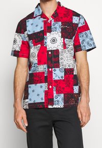 Camicetta a maniche corte con design a patchwork caratterizzata da colori rosso, blu, nero e bianco, e motivi paisley su tessuto di cotone.