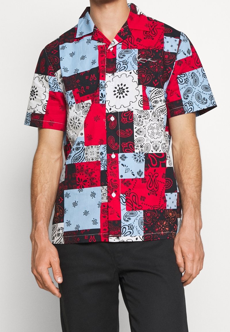 Camicetta a maniche corte con design a patchwork caratterizzata da colori rosso, blu, nero e bianco, e motivi paisley su tessuto di cotone.