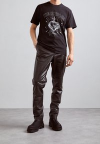 Camiseta gráfica negra con un diseño de corazón, combinada con pantalones de imitación de cuero negro brillantes y botas oscuras y robustas. Fondo simple.