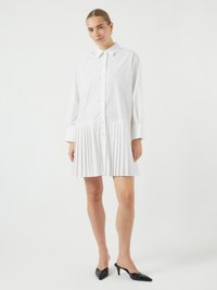 Robe chemise blanche à boutons sur le devant, manches longues et jupe plissée. Tissu lisse, col contrasté, portée avec des talons noirs.