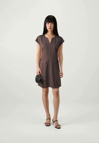 Brun kjole med cap-sleeves og V-hals, to frontlommer og buet kant; sammen med svarte stiletthæler og en liten svart håndveske.