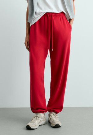 Persoon die rode joggingbroek met elastische boorden en beige sneakers draagt, staand tegen een lichtgrijze achtergrond.
