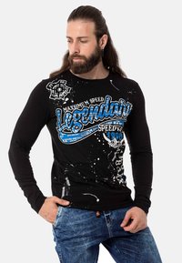 Schwarzes Langarm-T-Shirt mit blauem Grafiktext und Sprühfarben-Flecken. Kombiniert mit blauen, ausgewaschenen Jeans.