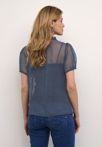 Blusa trasparente blu con maniche corte a palloncino, caratterizzata da un motivo testurizzato. Orlo arrotondato, abbinata a jeans blu. Vista posteriore mostrata.