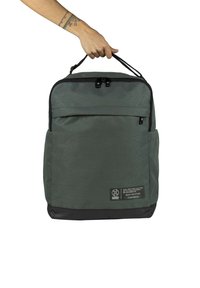 Mochila de tela verde con parte inferior negra, doble asa en la parte superior, bolsillo frontal con cremallera y parche con el logo en la esquina inferior. Material texturizado.