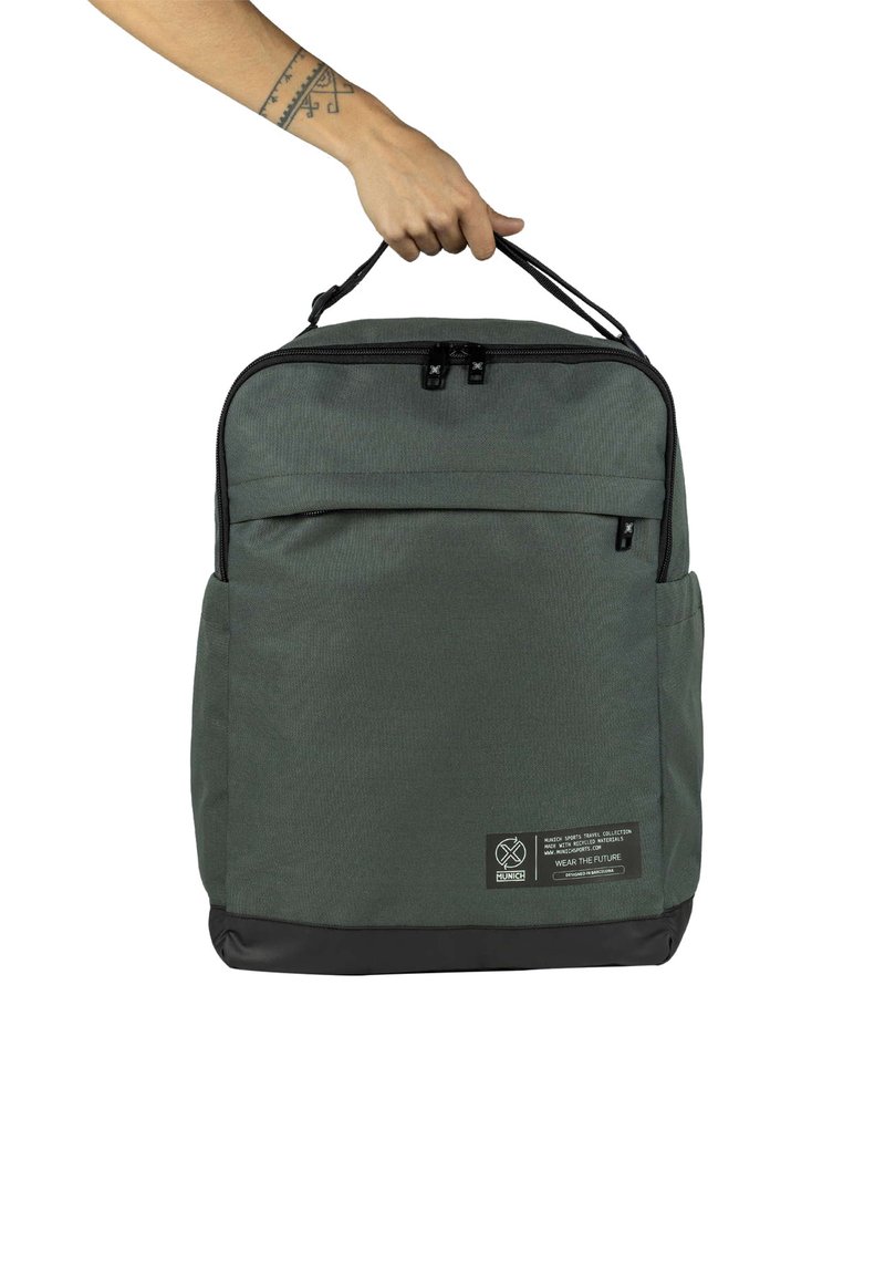 Mochila de tela verde con parte inferior negra, doble asa en la parte superior, bolsillo frontal con cremallera y parche con el logo en la esquina inferior. Material texturizado.