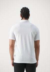 Vit kortärmad polo med klassisk krage, slät textur och minimalistisk design. Inkluderar sidofickor i fållen för ökad rörelsefrihet.