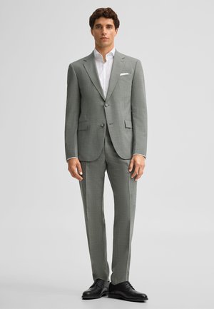 Homme debout portant un costume gris clair sur mesure avec une chemise blanche et des chaussures habillées noires, sur un fond clair uni.
