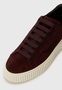 Baskets en daim marron foncé avec des lacets assortis et une épaisse semelle blanche striée, montrant des détails de face et de côté sur un fond blanc.