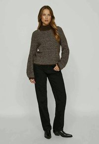 Brauner Heringenstrickpullover mit schwarzen Hosen; verfügt über einen gerippten Kragen und lange, strukturierte Ärmel, kombiniert mit Stiefeletten.