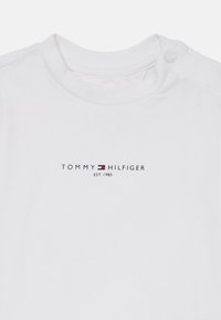 Weißer Baby-Body mit rundem Halsausschnitt und Druckknöpfen an der Schulter, mit dem Logo "Tommy Hilfiger EST. 1985" und Flagge in Schwarz, Rot und Weiß.