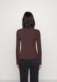 Långärmad topp i brun och rostfärgad randning med ribbad textur och vågig fåll, bärs med mörka jeans, sedd bakifrån.