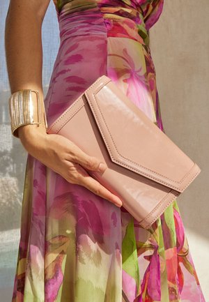 Mano che tiene una pochette envelope rosa chiaro, indossa un vestito floreale multicolore e un grande bracciale rigido dorato, su uno sfondo chiaro e neutro.