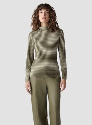 Woman with curly hair wearing an olive green and white striped turtleneck and olive green wide-leg pants against a plain background.