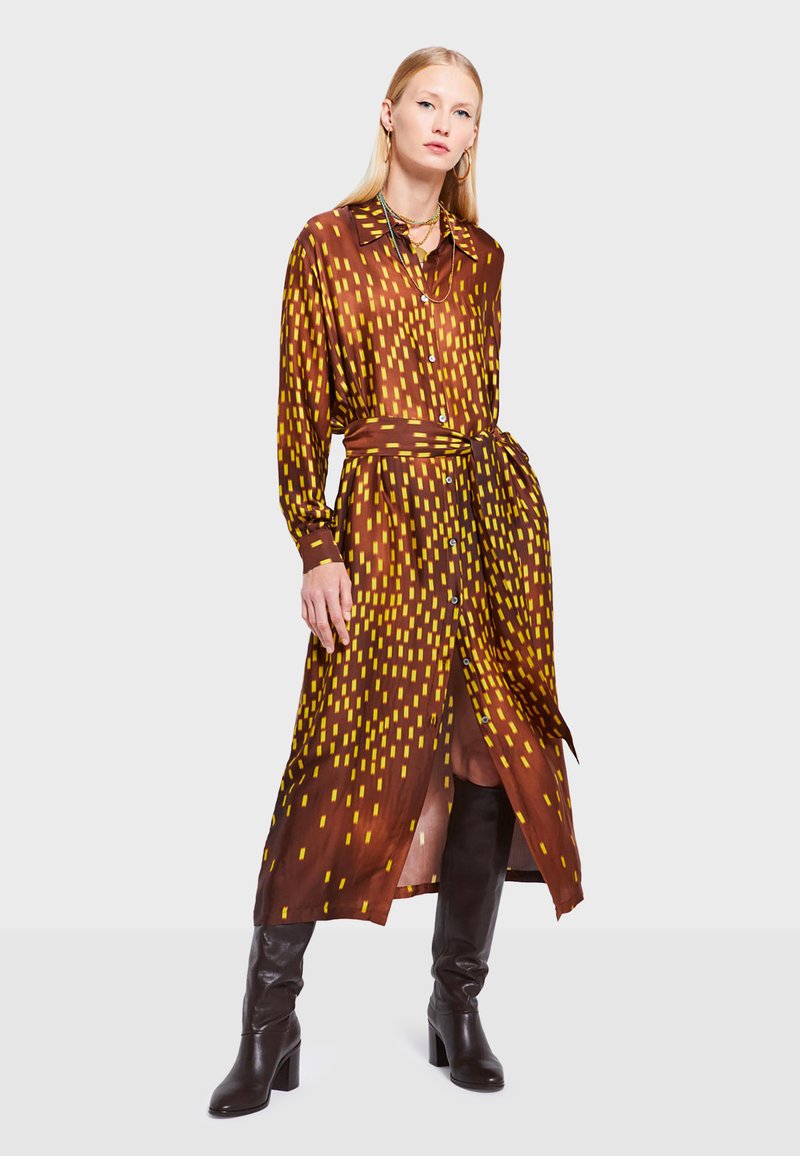 Abito lungo marrone con maniche lunghe e motivi geometrici gialli, chiusura a bottoni sul davanti, vita stretta con cintura e spacco alto, abbinato a stivali neri al ginocchio.