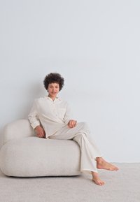 Lys beige loungewear med teksturert, ribbet mønster; løs skjorte med lomme og matchende bukser. Buet, myk kremfarget sofa.