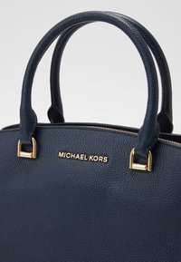 Sac à main en cuir texturé bleu foncé avec des accents métalliques dorés et des poignées courtes arrondies, avec le logo "MICHAEL KORS" en lettres dorées.