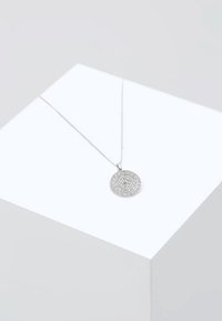 Collana d'argento con un pendente circolare che presenta un intricato motivo geometrico, appeso a una delicata catena, esposto su una superficie grigia chiara.