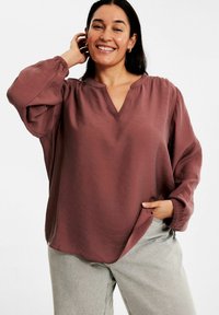 Kiansfärgad långärmad blus med V-ringning och rynkdetalj vid axlarna, tillverkad av lättviktigt tyg med en mjuk textur.