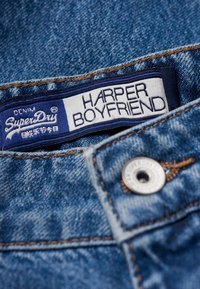 Tägning av denimtyg läser "SuperDry HARPER BOYFRIEND." Blå och vit med sömnadsdetalj; metallknappfäste i midjan.
