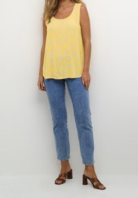 Geel mouwloos topje met een bloemmotief, gecombineerd met lichtblauwe rechtlopende jeans en bruine haksandalen.