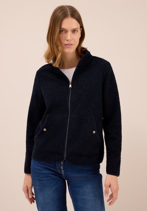 Veste en polaire bleu marine zippée avec un matériau texturé, deux poches avant et des boutons-pression argentés, portée sur un haut blanc.