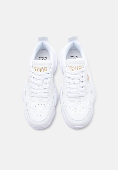 Paire de baskets basses blanches avec orteil perforé, lacets, et logo doré "Versace Jeans Couture" sur la languette et les côtés.