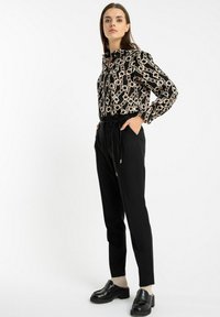 Kobieta stoi z rękami w kieszeniach, ubrana w czarne spodnie z wysokim stanem, czarne loafersy, beżowe skarpetki oraz czarną, wzorzystą bluzkę z długim rękawem.