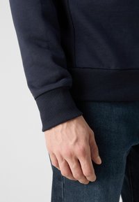 Hanorac bleumarin cu manșete și tiv cu model de rigide; amestec de bumbac, textură moale; mână odihnindu-se pe țesătură de denim, arătând un detaliu în apropiere.