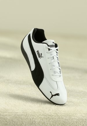 Baskets Puma blanches et noires avec logo sur le côté et la pointe, présentées en angle sur une surface beige texturée.