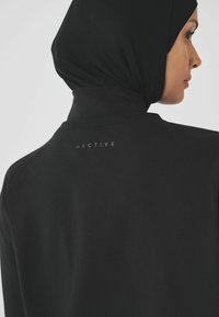 Melnā sportiskā augšdaļa ar augstu apkakli, maigu tekstūru un bezšuvju dizainu. "ACTIVE" logo kontrastējošā drukā muguras apkakles vietā.
