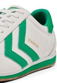 Sapatilha branca com acidentes verdes, apresentando um cabedal sintético texturizado, costuras em contraste, e o logo dourado "STADION" na lateral.