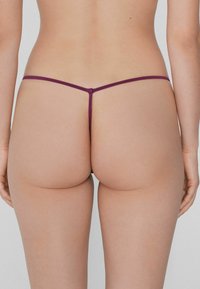 String violet en dentelle avec des bretelles fines et une couverture minimale, mettant en valeur une texture de peau lisse et nude. Le design élégant souligne la simplicité.