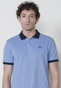 Gaiši zils polo krekls ar tekstūrētu rakstu, tumši zila apkakle un piedurkņu maliņas, un trīs pogu aizdare. Iekļauj izšūtu logo uz krūtīm.