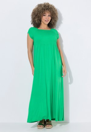 Vrouw met krullend haar, gekleed in een felgroene maxi-jurk met korte mouwen en zwarte platformsandalen, staand tegen een witte achtergrond.