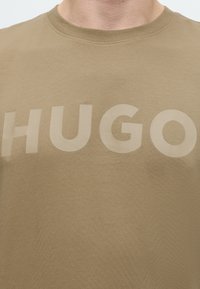 Κοντινό πλάνο ενός ατόμου που φοράει μπεζ t-shirt με στρογγυλή λαιμόκοψη και μεγάλο, έντονο κείμενο "HUGO" τυπωμένο στο στήθος.