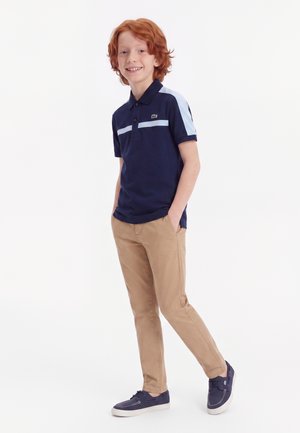Garçon roux souriant, debout les mains dans les poches, portant un polo bleu marine, un pantalon beige et des chaussures décontractées bleu marine sur fond blanc.